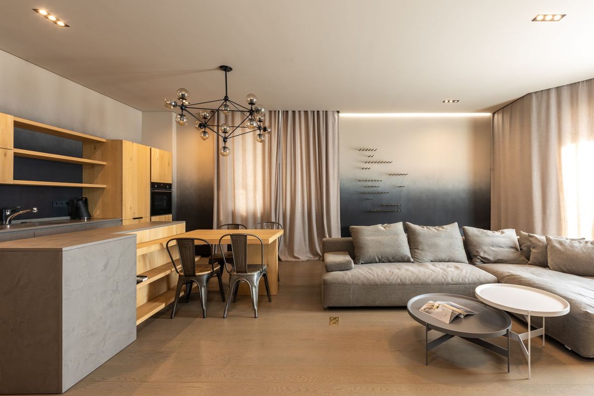 Comfortable couch with cushions near coffee table placed near kitchen with wooden cupboards in light modern apartment with curtains and windows