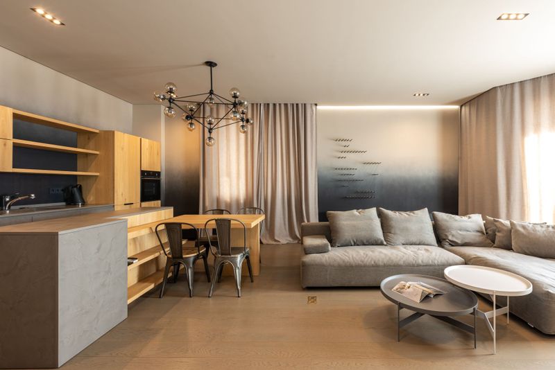 Comfortable couch with cushions near coffee table placed near kitchen with wooden cupboards in light modern apartment with curtains and windows