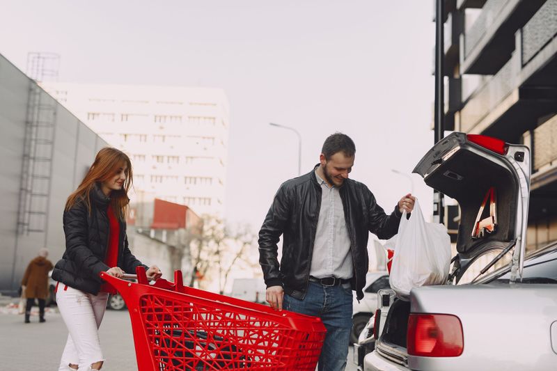 From below of smiling couple in casual clothes moving shopping bags from supermarket cart to car trunk on street parking